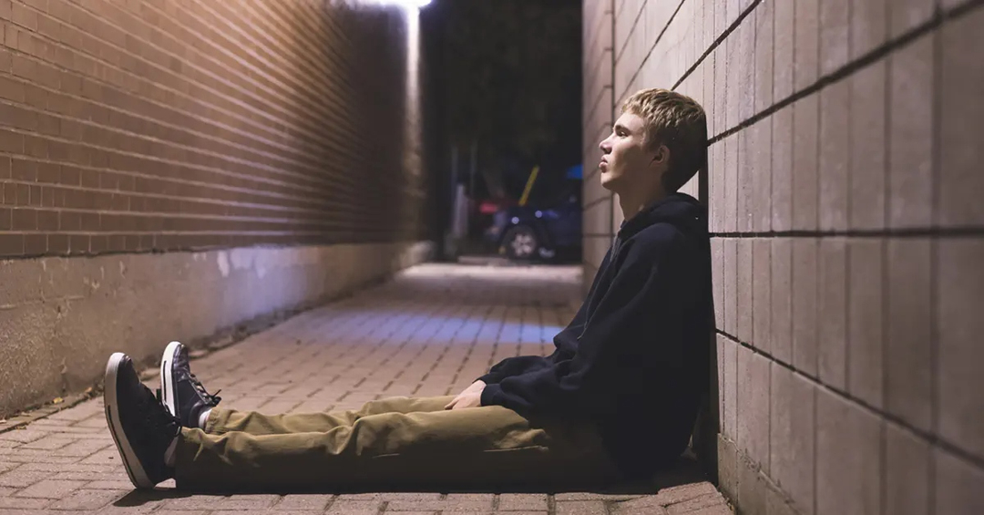 Young adult sitting in an alley at night leaning against a wall