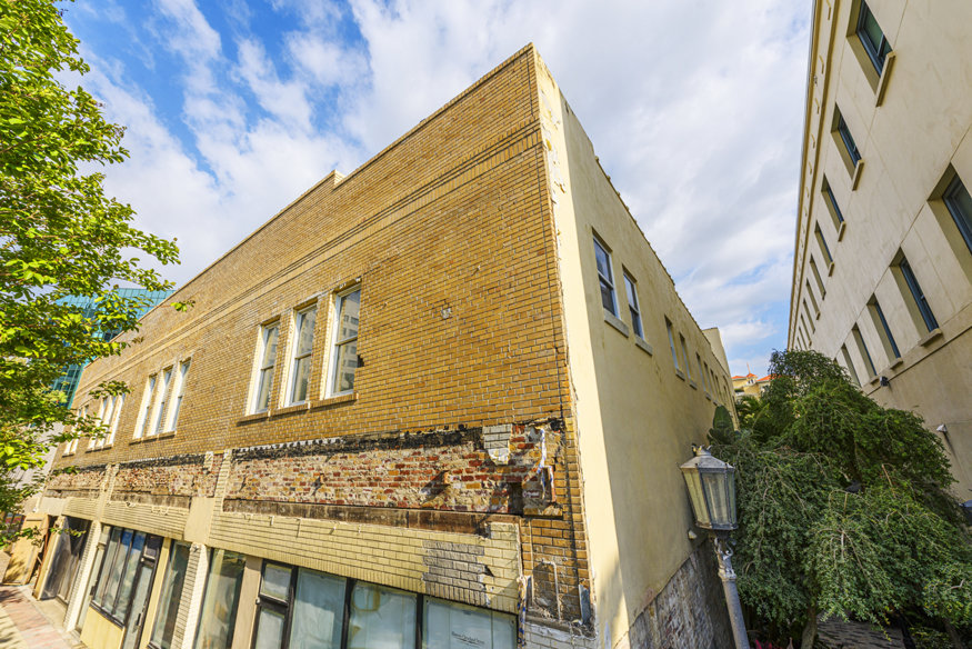 A two-story brick building with parts of its façade uncovered during restoration work, revealing historic 1928 masonry beneath layers of paint and nonoriginal materials. Adjacent buildings and a leafy tree are visible under a partly cloudy sky.
