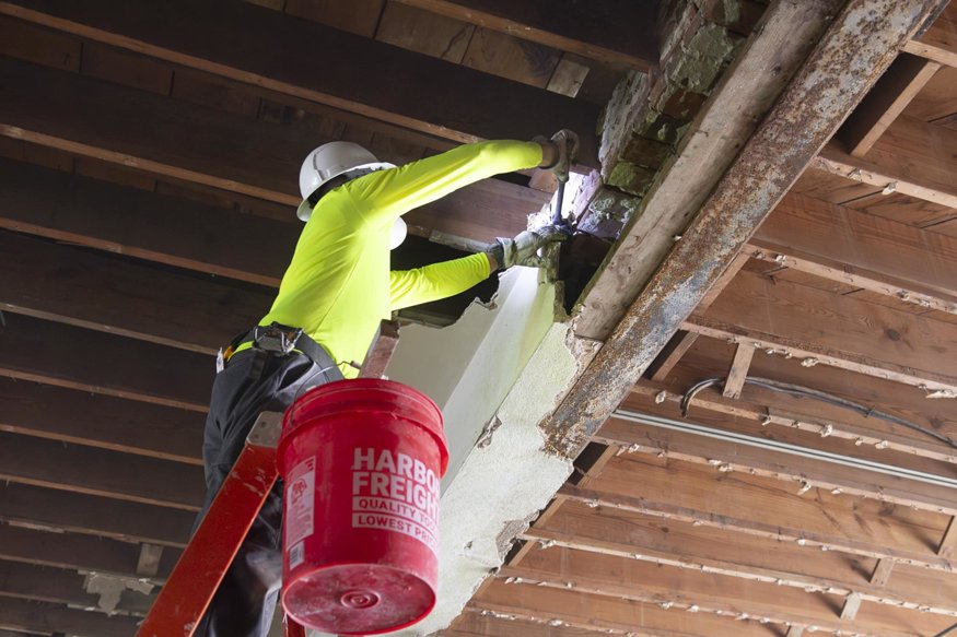 Worker uses hammer to remove plaster near structural beam.