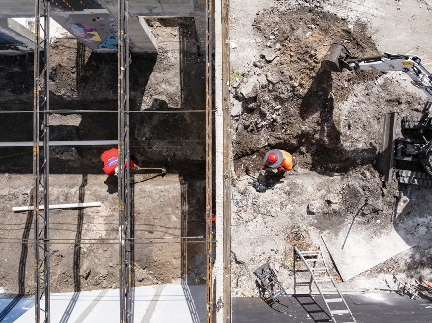 Workers prepare plumbing trenches behind building.