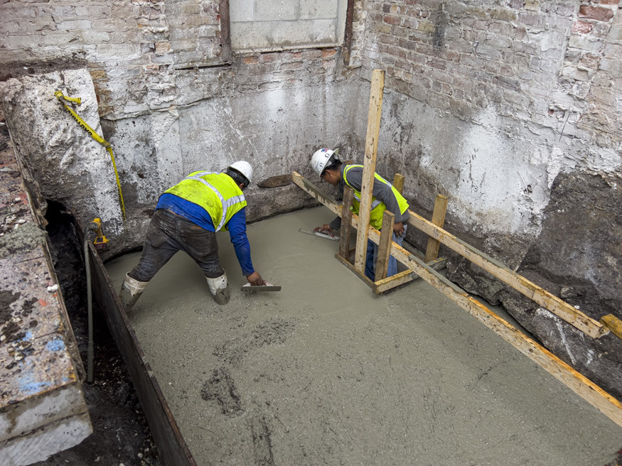 Workers smooth and level concrete at the bottom of the elevator shaft.