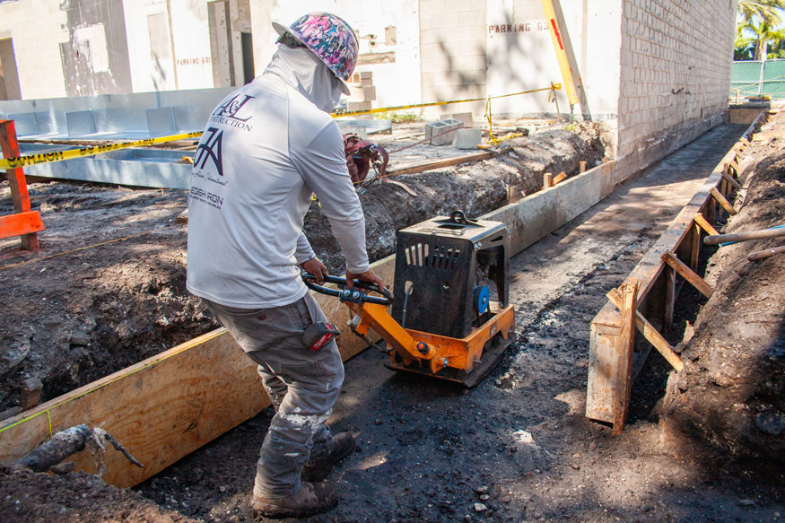 Worker operates plate compactor in footing area.
