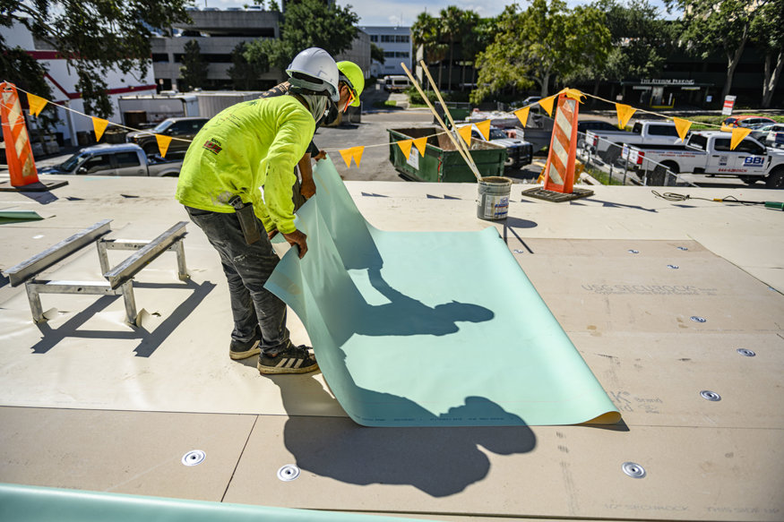 Workers position roofing membrane across roof surface.