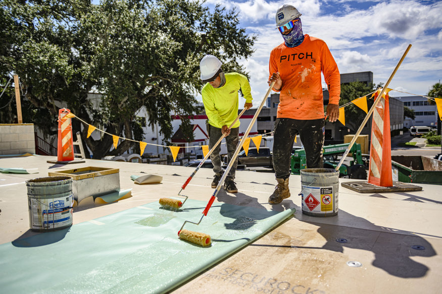 Workers apply bonding material to roofing membrane.