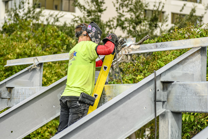 Welder prepares steel framing for metal decking along sloped soffit.