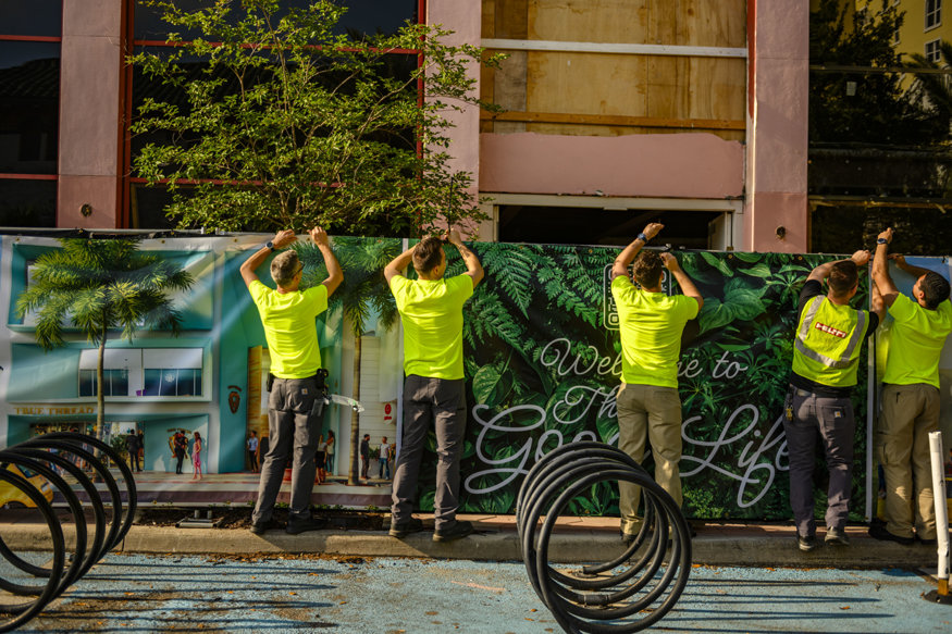 Workers align construction panels along sidewalk fencing. 