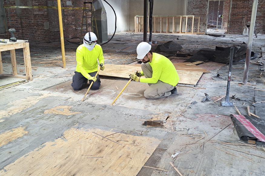wo construction workers in bright yellow shirts and hard hats using tools to remove old flooring inside the historic Peoples Bank building during the early stages of its restoration.