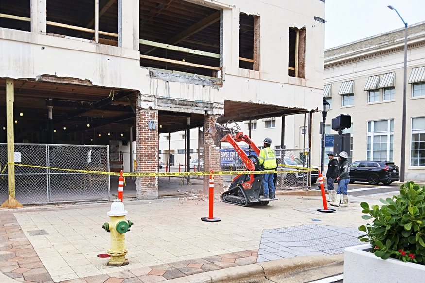 A construction site at the Peoples Bank building with workers using equipment to demolish remaining sections of the façade, while new concrete walls begin to take shape. Safety cones and tape surround the area.