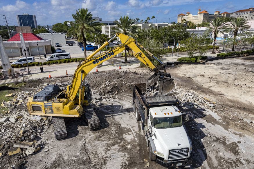 Excavator loads concrete debris into truck on cleared lot. 
