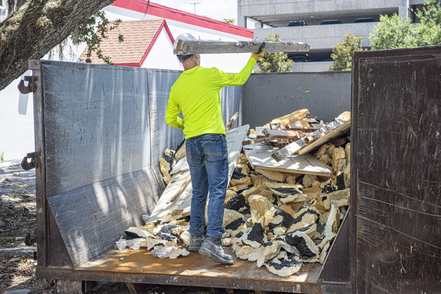 Worker unloads wall debris into a large dumpster outside.