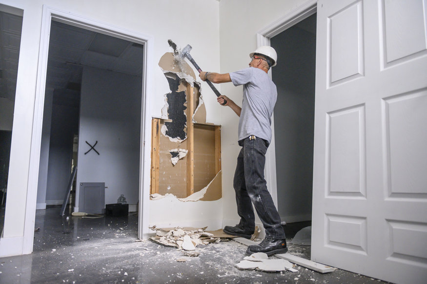 Construction worker removes drywall to expose wood framing and black insulation.