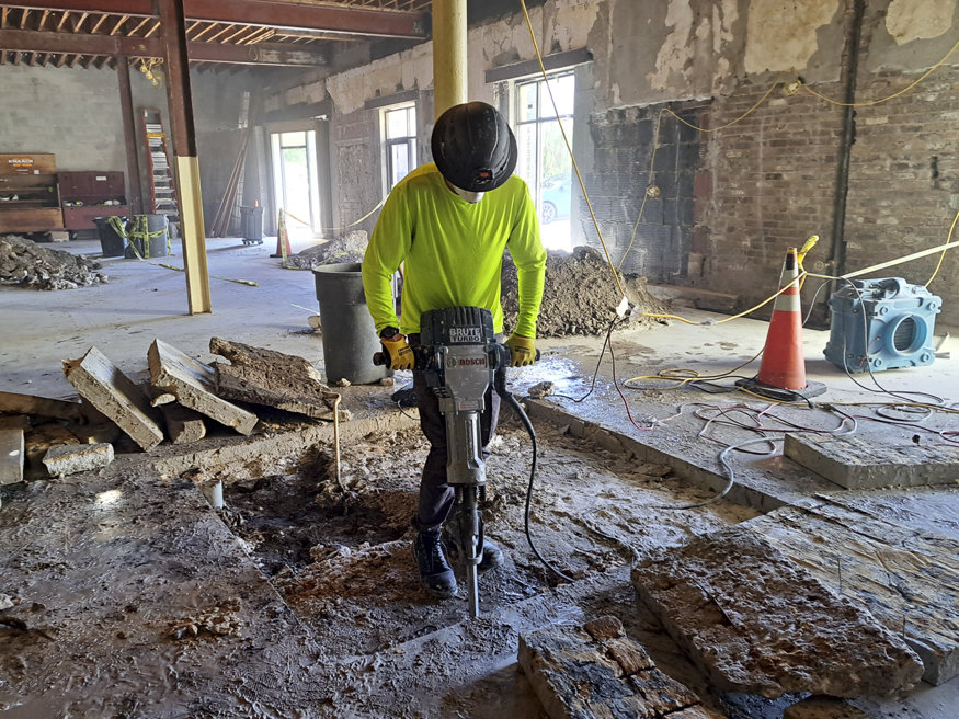 A worker in a hard hat and neon shirt operates a jackhammer inside a worn historic building, breaking up old concrete as part of the restoration effort to reinforce the foundation and floors.