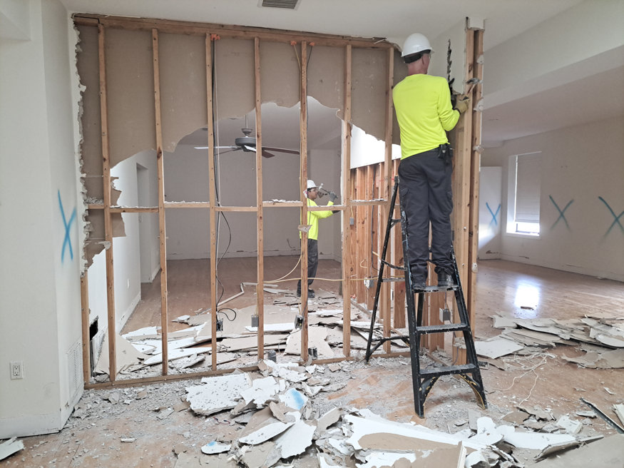 Workers remove drywall and outdated materials from a wall, revealing wooden studs as part of a full interior demolition process.