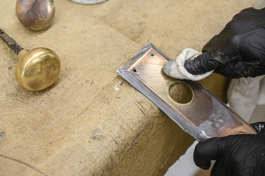 Gloved hands clean an antique metal door plate.