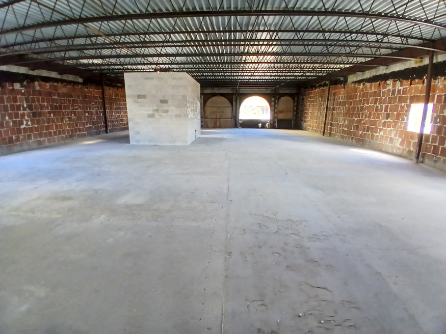 An empty, open space inside a brick and steel framework with a central block structure under a metal roof.