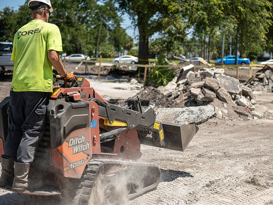 Construction worker operating a compact track loader carrying broken concrete during foundation removal.