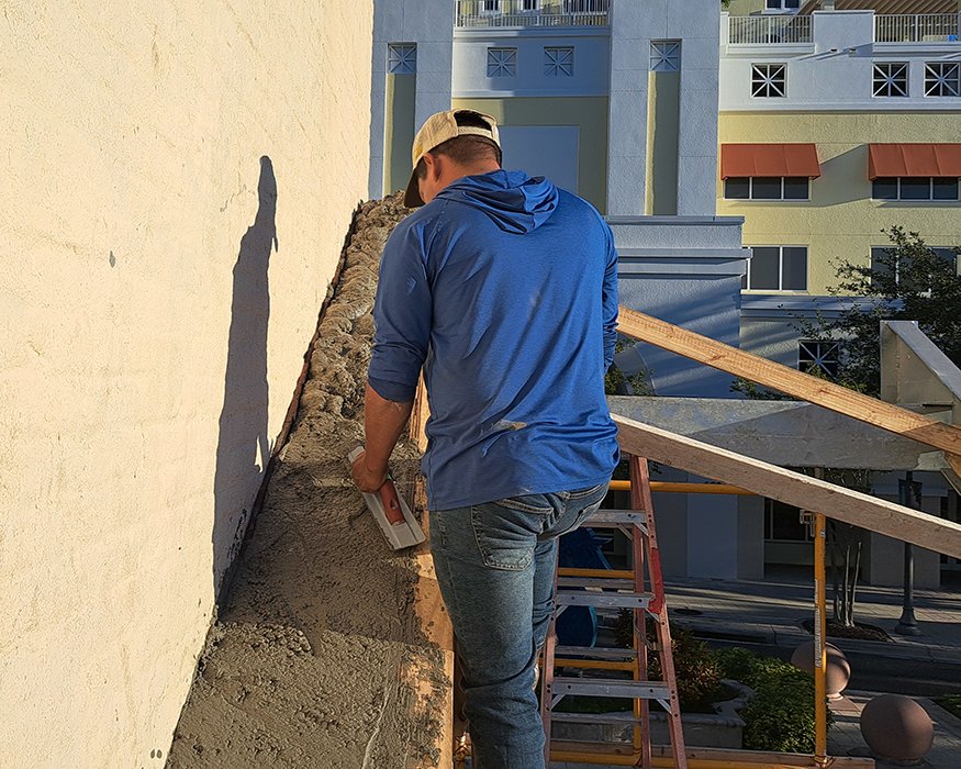 Worker smooths concrete along sloped beam beside building wall.