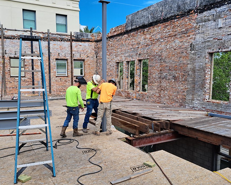 Three workers guide steel column into place by open joist bay.