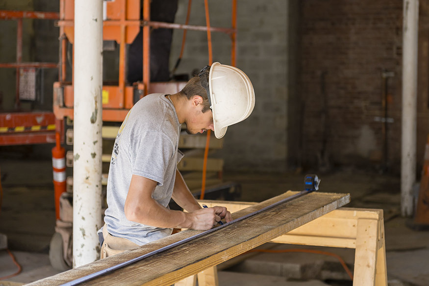 Worker marking cut lines on a wooden beam at a sawhorse inside renovation site.