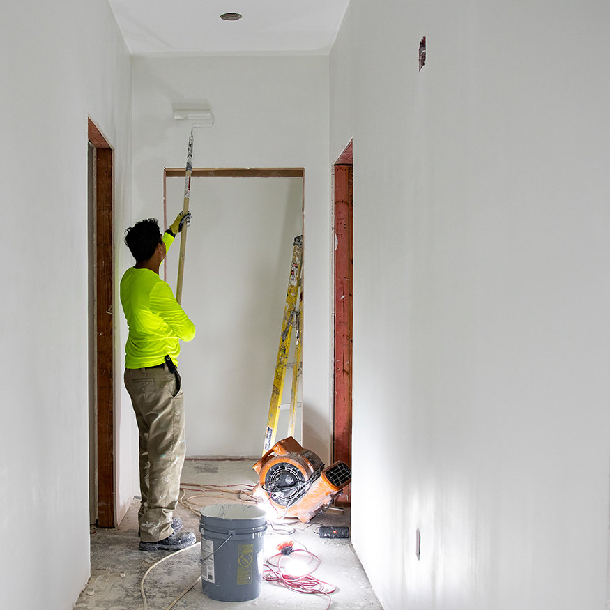 Painter using extended roller beneath tall interior ceiling. 