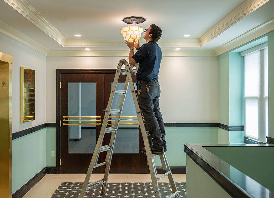 Worker standing on ladder installing gold-accented multi-layered flush-mounted ceiling light.