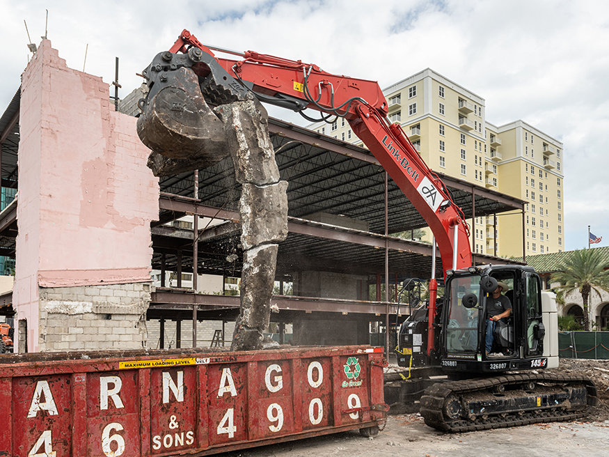 Excavator places concrete footing debris into dumpster during demolition.