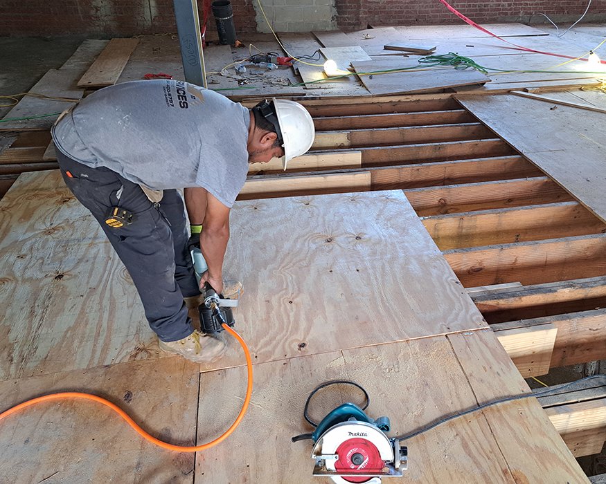 Worker nailing plywood floor decking over open joists at upper level.