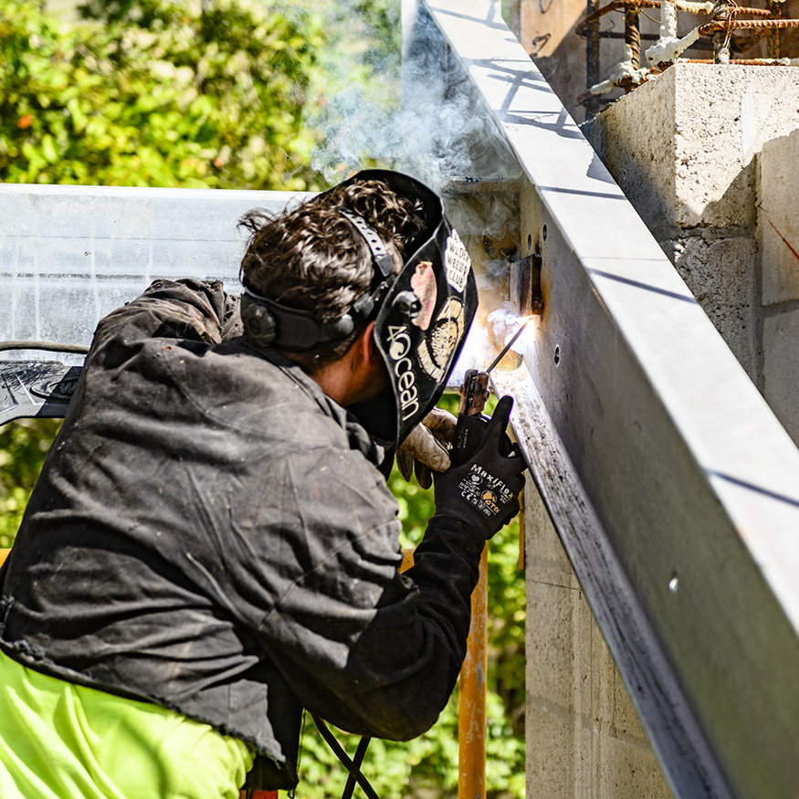 Worker welds steel connection at sloped entry framing.