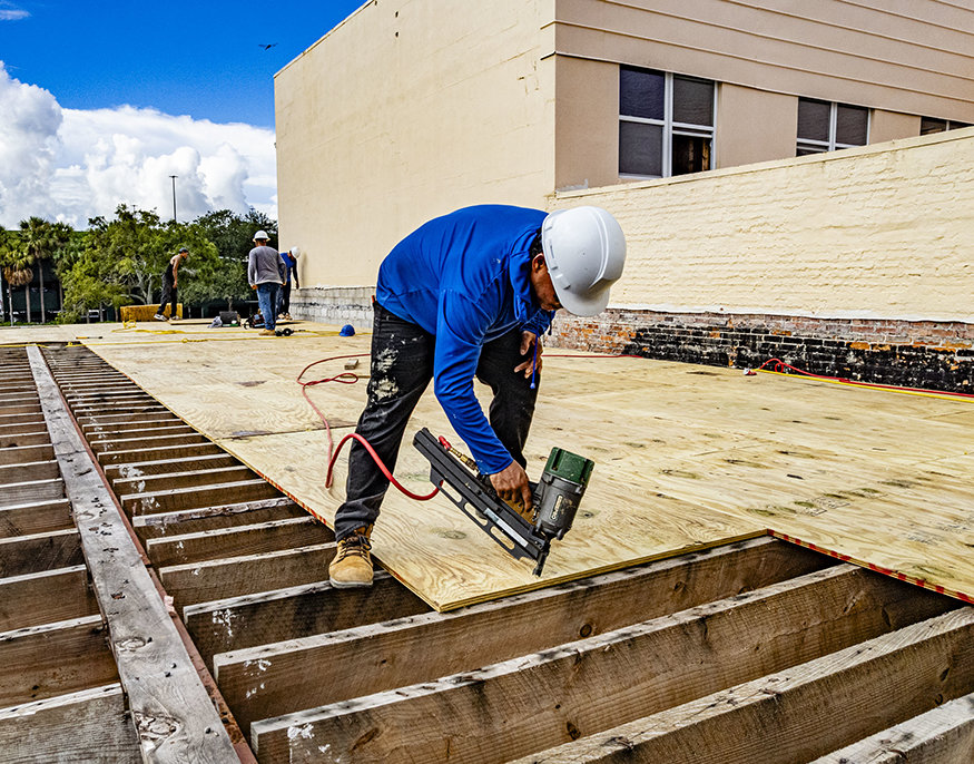 Worker fastens plywood decking with pneumatic nailer.