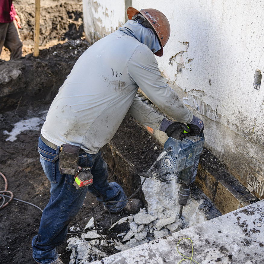Worker uses demolition hammer along footing excavation.