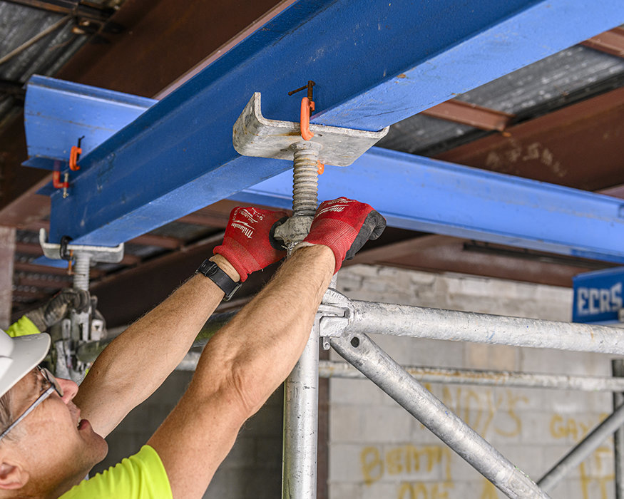 Worker adjusts overhead shoring jack supporting structural steel.